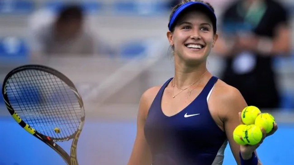 Eugénie Bouchard tout sourire sur un terrain de tennis, elle a quatre balles jaune dans sa main gauche et une raquette dans l'autre.
