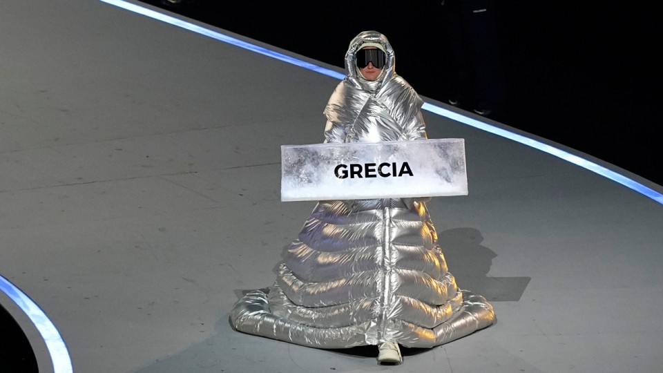 Une personne marche sur l'allée des Jeux Olympiques en portant un signe où il est écrit Grèce. Elle porte un manteau long d'un argent éblouissant, ainsi que de grosses lunettes de ski.