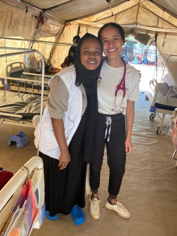 On voit deux docteures côte-à-côte. Derrière elles, il y a plusieurs lits d'hôpital.  Elles sont dans une grande tente. 