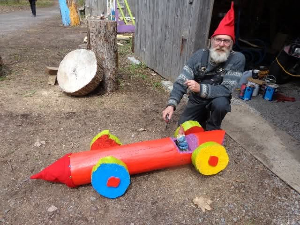 On voit un homme avec des lunettes et une grosse barbe. Il porte un chapeau de gnome et a un genou par terre. Il est à côté d'une voiture de bois que conduit une petite figurine de gnome.