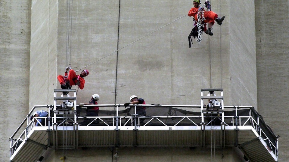 On voit deux hommes qui sont en train de grimper avec des cordes le long d'une tour. Ils sont habillés en orange, avec des casques. Proche d'eux, deux autres personnes qui portent aussi des casques s'assurent que ça se passe bien. 